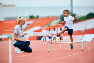 Sports,Teacher,Using,Stopwatch,While,Boy,Is,Jumping,Over,Hurdles